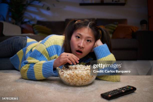 young asian girl lying on the living room floor watching a movie at home, amazed by what's on tv. - escandalo tv fotografías e imágenes de stock