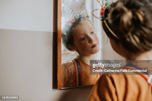 a young girl wearing a quirky unicorn headband and rainbow braces looks at her reflection in a mirror - kopf zur seite neigen stock-fotos und bilder