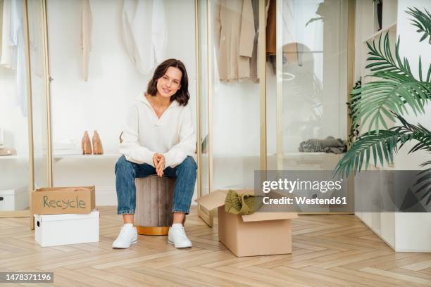 happy young woman sitting on stool and recycling clothes at home - de clutter stock pictures, royalty-free photos & images