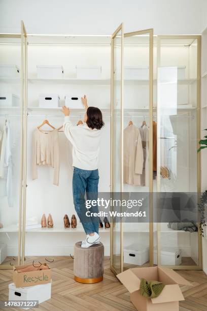 young woman standing on stool and removing box from closet at home - quehaceres-domésticos fotografías e imágenes de stock