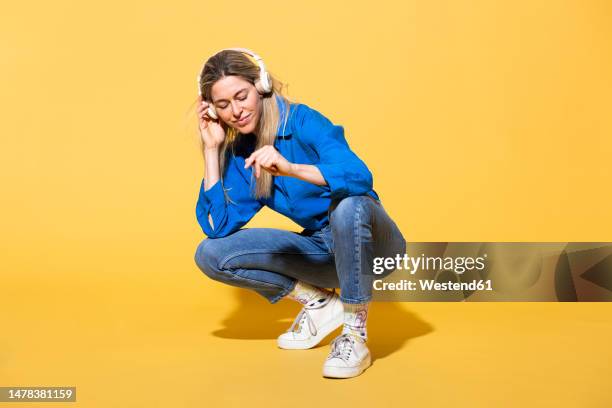 woman listening to music and crouching against yellow background - frau kopfhörer indoor stock-fotos und bilder