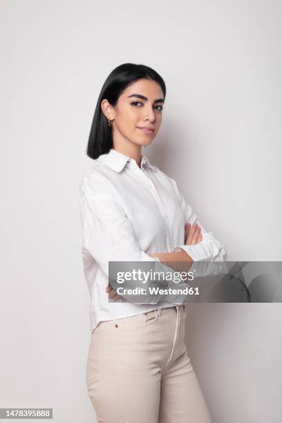 confident young woman standing with arms crossed against white background - blusa camisas fotografías e imágenes de stock