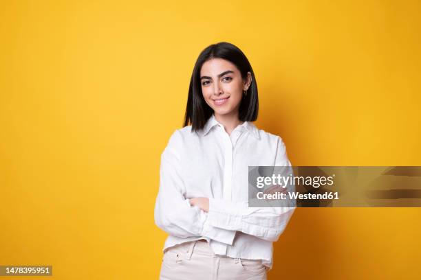 smiling young woman standing with arms crossed against yellow background - arms crossed fotografías e imágenes de stock