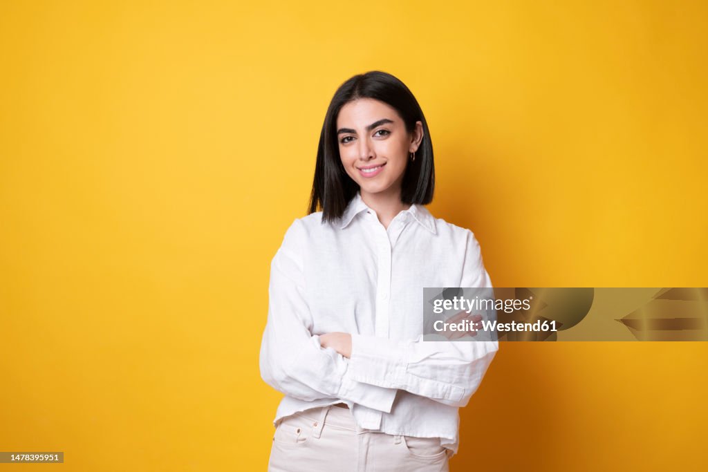 Smiling young woman standing with arms crossed against yellow background