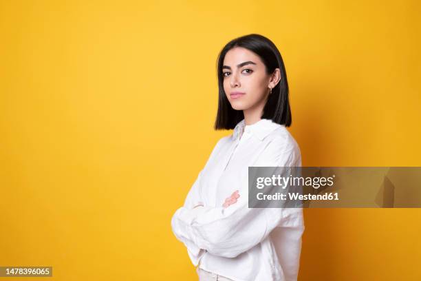 young woman standing with arms crossed against yellow background - fondo amarillo fotografías e imágenes de stock