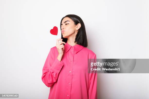 young woman puckering at red heart shaped lollipop against white background - young at heart ストックフォトと画像