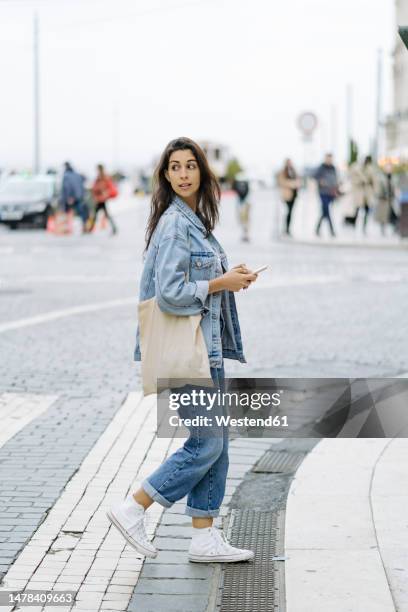 young woman with tote bag and smart phone crossing road - mirar por encima del hombro fotografías e imágenes de stock