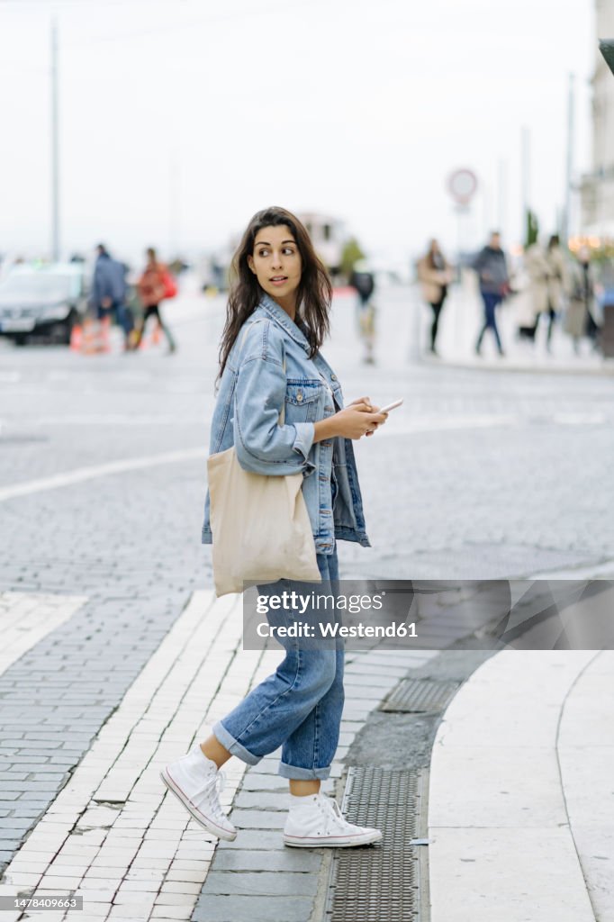 Young woman with tote bag and smart phone crossing road