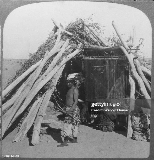 Navajo man stands wearing a war bonnet, holding a bow and arrows, as a woman behind him weaves a blanket in a traditional Navajo hogan on the Navajo...