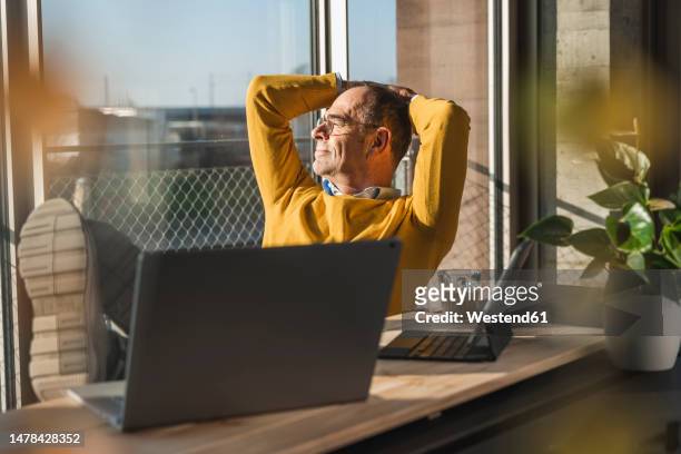 contemplative businessman sitting with hands behind head at desk in office - kleine scherptediepte stockfoto's en -beelden