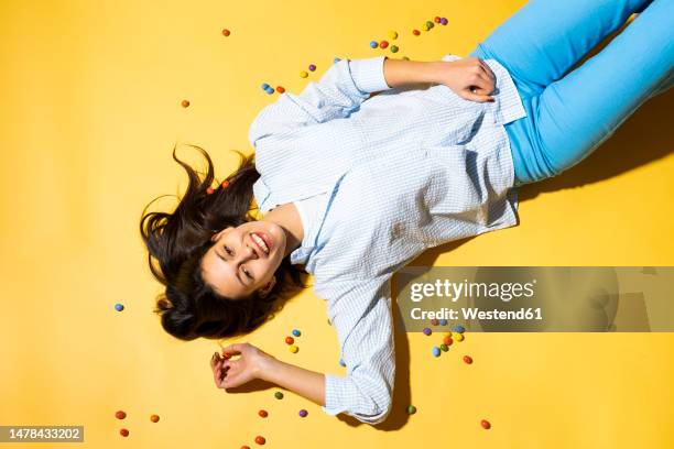 happy young woman lying amidst colorful candies yellow background - supino foto e immagini stock