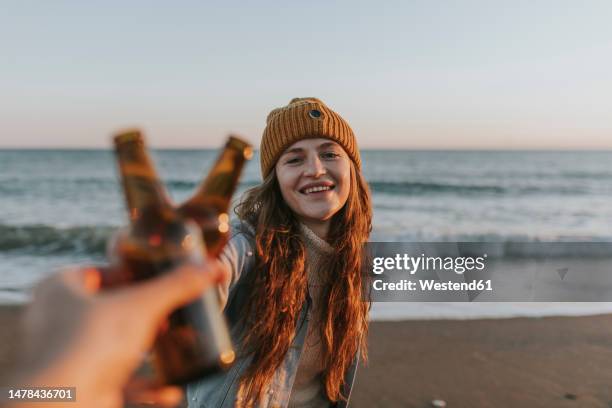 happy woman toasting beer bottle with friend in front of sea - beer bottle fotografías e imágenes de stock