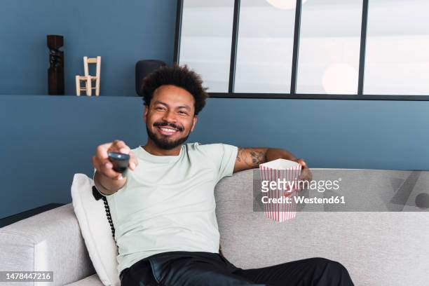 smiling man watching tv sitting on sofa at home - changing channels stockfoto's en -beelden