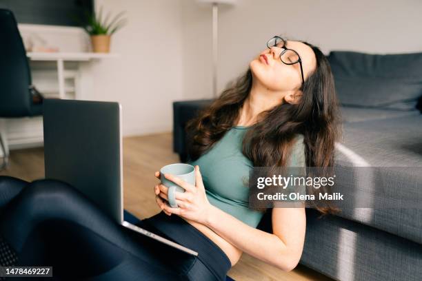 young brunette woman with coffee cup and laptop is tired while working from home - millenials stock-fotos und bilder