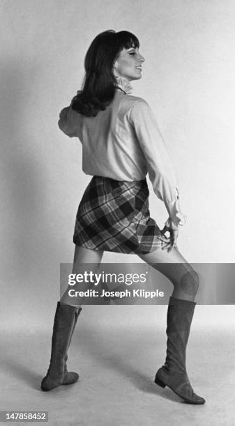 Profile portrait of an unidentified fashion model dressed wearing a long-sleeve shirt with a ruffled collar, a plaid mini-skirt, and low-heel, suede...
