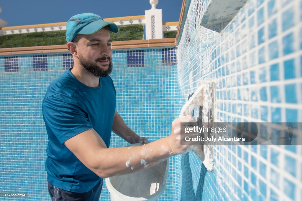 Bearded man spreads the cement grout on the pool tile to waterproof it, preparing pool for summer