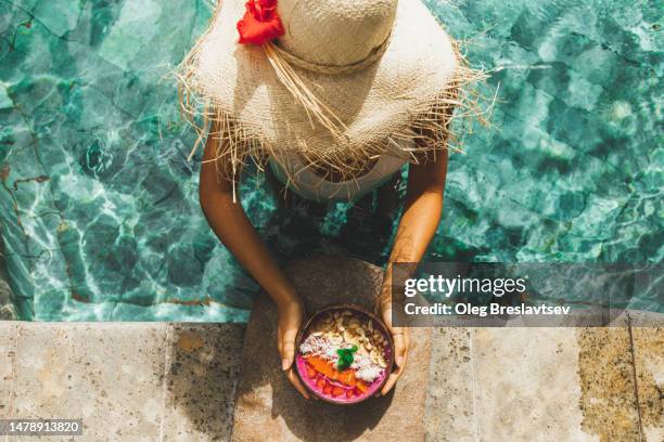 overhead view of woman in straw hat holding smoothie bowl on poolside.  emerald clear water in pool - bali luxury stock pictures, royalty-free photos & images