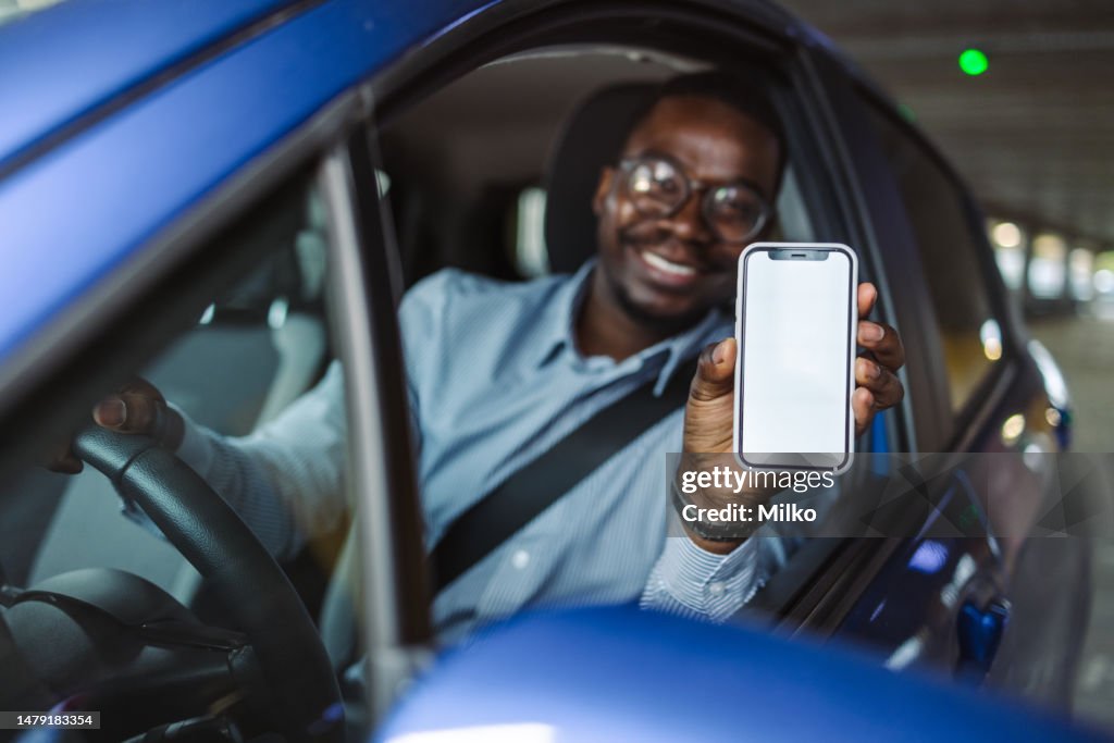 Cheerful African-American man sitting in his car in a parking garage and showing a smart phone with a blank screen