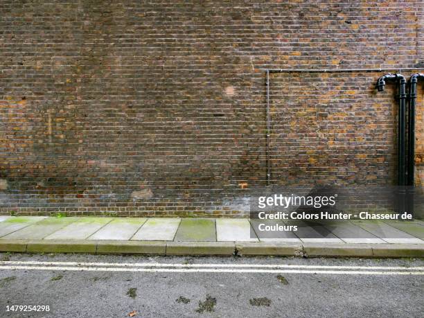 weathered brown brick wall with black pipes, concrete sidewalk and asphalt street with markings in london, england, united kingdom - dachgiebel stock-fotos und bilder