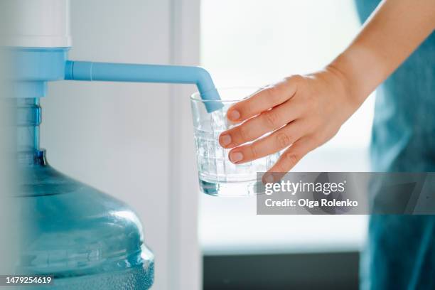 cropped human hand pouring fresh mineral water from water cooler to drinking glass against bright window - filtered foto e immagini stock