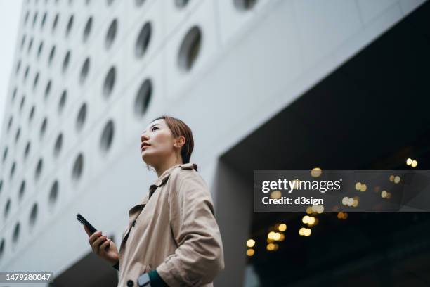 low angle portrait of a successful young asian businesswoman with smartphone in the city, standing against urban corporate skyscrapers in downtown area. looking up and aiming high. woman entrepreneur in business. business success and achievement - access foto e immagini stock