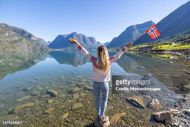 woman holds norwegian flag against lake and mountain landscape - norwegian culture stockfoto's en -beelden