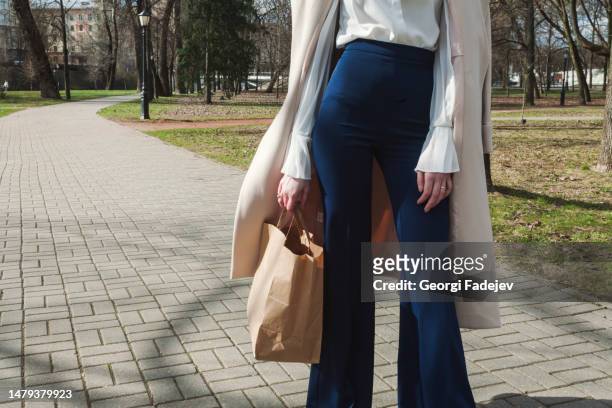 fashionable unrecognizable girl in the park, with a coat thrown over her shoulders, smart watch on her wrist, stylish tight dark blue pants, white shirt. holds paper bag. - pantaloni a zampa di elefante foto e immagini stock