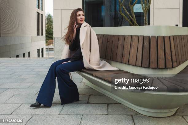 a beautiful businesswoman with a white coat draped over her shoulders sits on a bench in the business district with a phone in her hand. - bootcut pants stockfoto's en -beelden