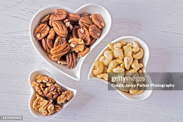 close-up, still-life, flatlay image of a collection of white heart-shaped bowls filled with mixed nuts, pecans, cashews and walnuts - cashewnuss stock-fotos und bilder