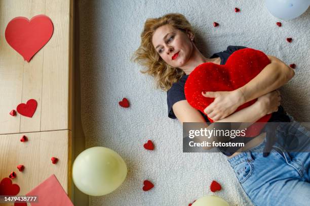 woman in love lying on the floor, embracing a heart shaped pillow and daydreaming - verliefd worden stockfoto's en -beelden