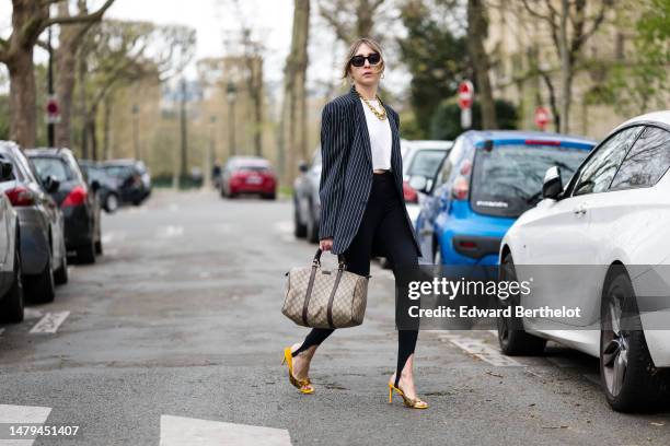 Emy Venturini wears black squared sunglasses, gold earrings, a large gold necklace, a white t-shirt, a black with white striped print pattern...