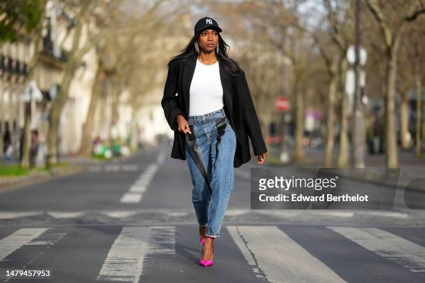 Emilie Joseph wears a black with embroidered white NY logo pattern cap, crystals fringed pendant earrings, a white t-shirt, a black oversized blazer...