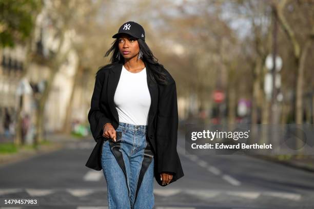 Emilie Joseph wears a black with embroidered white NY logo pattern cap, crystals fringed pendant earrings, a white t-shirt, a black oversized blazer...