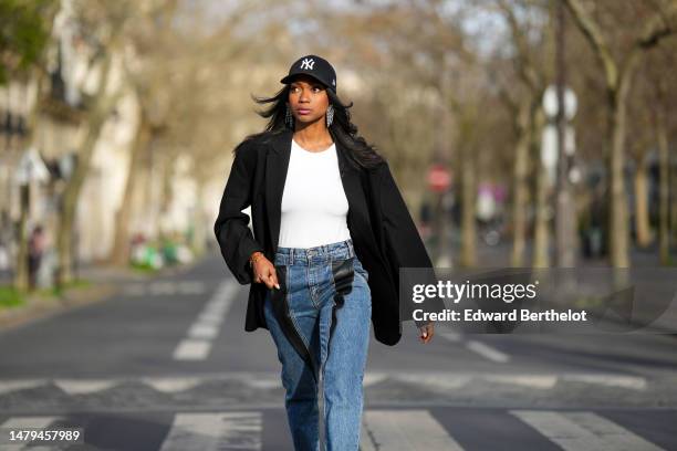 Emilie Joseph wears a black with embroidered white NY logo pattern cap, crystals fringed pendant earrings, a white t-shirt, a black oversized blazer...