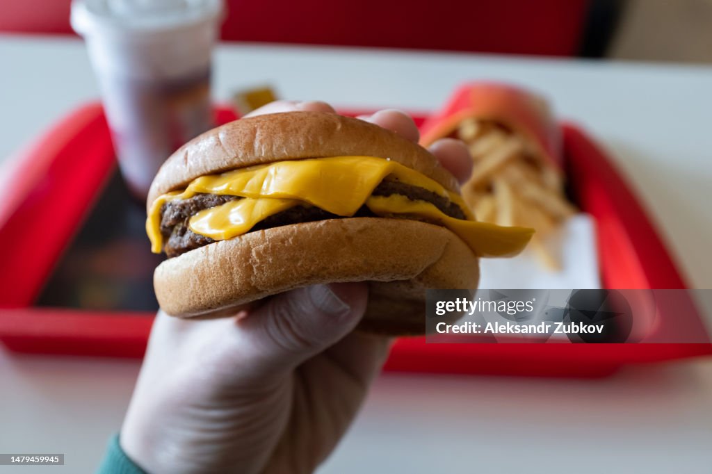 A young man is holding a piece of hamburger in his hands. A guy or a man eats fast food. A hungry skinny guy is eating an appetizing burger. The concept of unhealthy food, diet, overeating, gluttony, dependence on food. Fast food restaurant, snack bar.