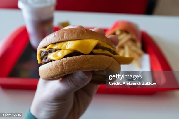 a young man is holding a piece of hamburger in his hands. a guy or a man eats fast food. a hungry skinny guy is eating an appetizing burger. the concept of unhealthy food, diet, overeating, gluttony, dependence on food. fast food restaurant, snack bar. - snabbmat bildbanksfoton och bilder