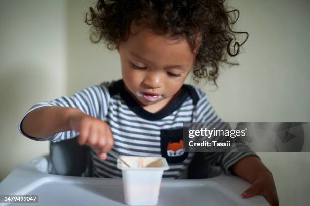 snack, dairy and child eating yoghurt in his high chair for hunger, healthy craving or dessert. hungry, food and little boy kid or toddler enjoying a yogurt for milk nutrition with a spoon at home. - baby eating yogurt stockfoto's en -beelden