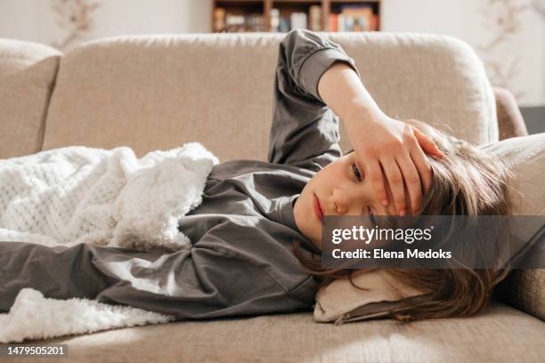 a sick dark-haired teenage girl lies on the sofa, covered with a blanket and put her hand on her forehead - headache child fotografías e imágenes de stock