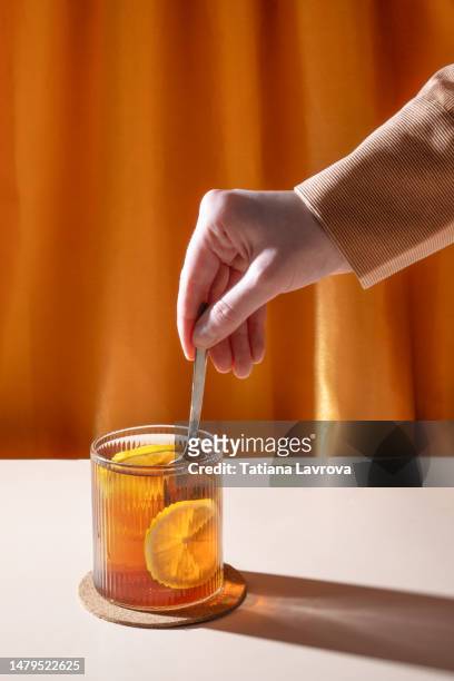 hand making tea with lemons by putting tea bag into transparent glass against orange curtains background with beautiful illuminated shadow.  closeup of natural hot beverage. relaxation, wellness, self-care concept - chamomile tea bag stock pictures, royalty-free photos & images