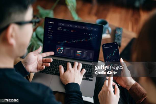 young asian couple managing finance and investment online, analyzing stock market trades with mobile app on laptop and smartphone. making financial plans. banking and finance, investment, financial trading, mobile banking concept - accesorio financiero fotografías e imágenes de stock
