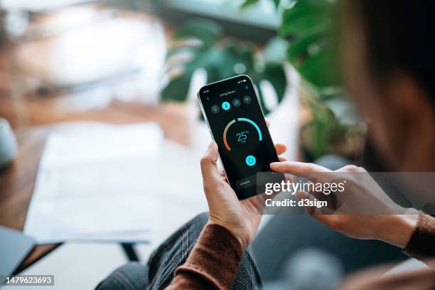 over the shoulder view of asian woman holding smartphone, setting up the intelligent home system, remote-controlling the thermostat of smart air-conditioner in the living room of her smart home. smart living. home automation. lifestyle and technology - intelligenz stock-fotos und bilder
