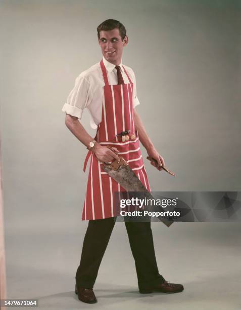 Posed studio portrait of a male fashion model wearing a red and white striped apron over a white shirt and tie, he carries tools in the front pocket...