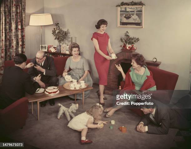 House party scene with adults and a child seated in a living room being served cups of tea and cake by a woman hosting the occasion, London, 27th...