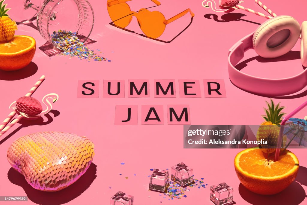Flat lay of bright colored oranges served as cocktail, kawaii headphones, cocktail glass filled with confetti, heart shaped sunglasses, ice, and text Summer jam on soft pink background