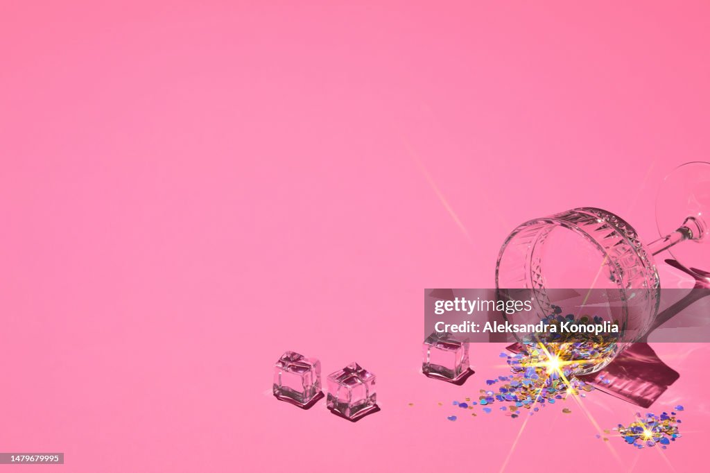 Summer flat lay - ice, crystal champagne cocktail glass filled with holographic heart shaped confetti with starry glares on soft pink background with copy space
