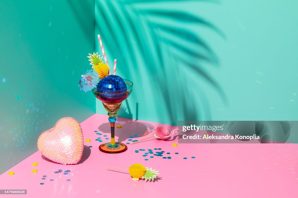 Still life scene with composition of colorful cocktail glass served as ice cream with blue sparkling disco ball and cute pineapple paper straws, heart with sequins, confetti and tropical palm tree leaf shadow on solid pink and mint green background