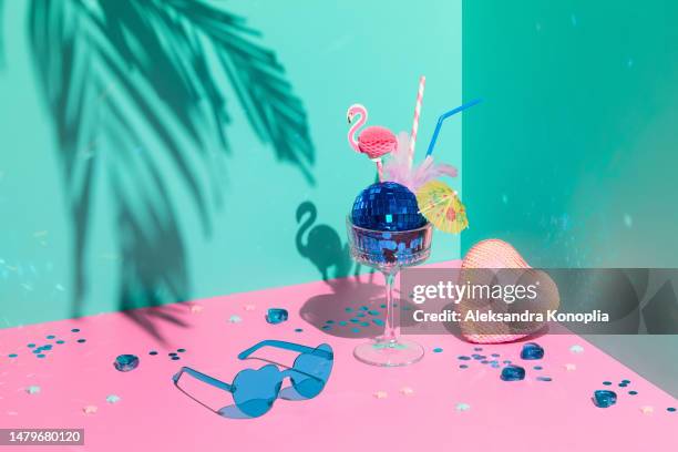 still life scene with composition of colorful cocktail glass served as ice cream with blue sparkling disco ball and cute pineapple paper straws, heart with sequins, confetti and tropical palm tree leaf shadow on solid pink and mint green background - flamingo heart fotografías e imágenes de stock