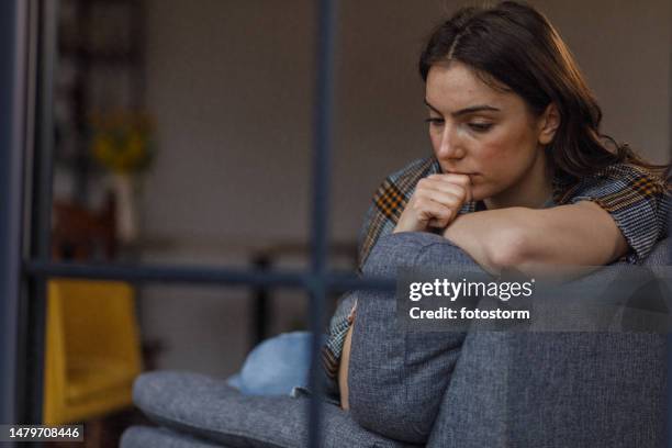sad young woman sitting on the sofa, wrapped in a blanket, contemplating problems and solutions - nail biting stock pictures, royalty-free photos & images