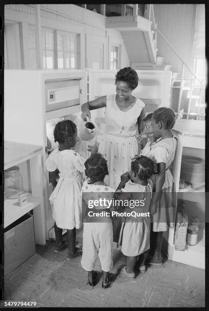 Group of child cluster around a woman who smiles as she pours them drinks from a pitcher, Little Rock, Arkansas, August 20, 1959.