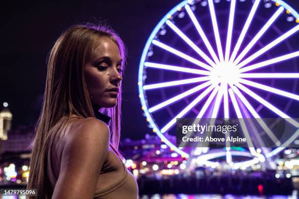 bella donna bionda al festival in piedi di fronte alla grande ruota di seattle di notte luci della città sullo sfondo la ruota panoramica illuminata lunga esposizione - woman capturing city night foto e immagini stock